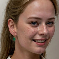 Gold drop earrings with emerald-green oval stones, worn by a smiling model