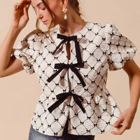 Ivory woven peplum top with black bow tie at neckline and puff sleeves, angled side view.