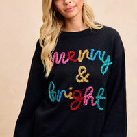 Black holiday sweater with multicolor tinsel "Merry & Bright" lettering at center front