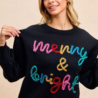 Black sweater featuring multicolor tinsel "Merry & Bright" text across the front