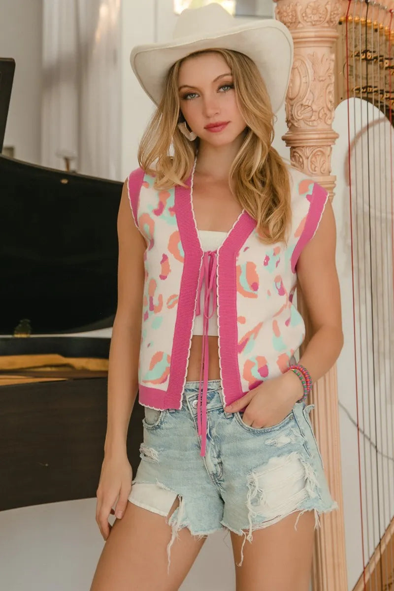 Sleeveless ivory sweater vest with pink leopard-style print, V-neck tie cords, and scalloped hem.