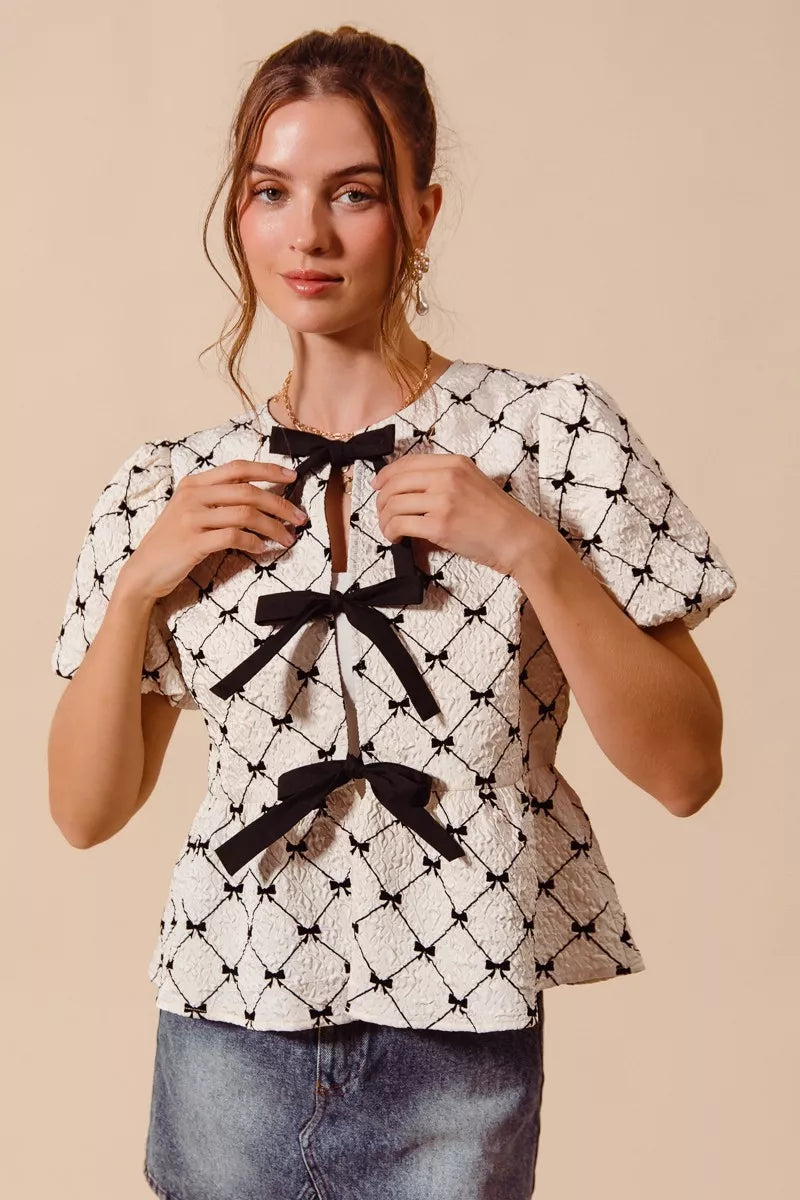 Ivory woven peplum top with black bow tie at neckline and puff sleeves, front view.