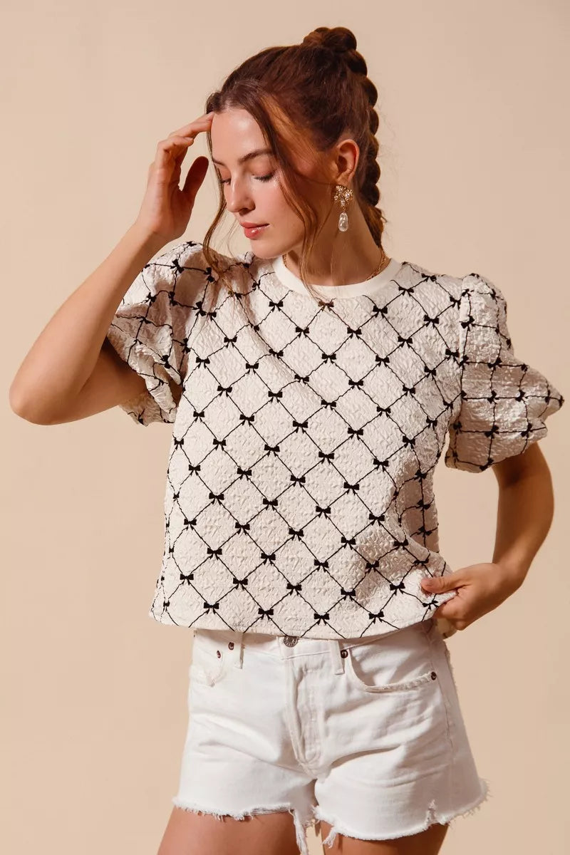 Ivory woven bubble sleeve top with black bow texture pattern, hands-to-ponytail pose