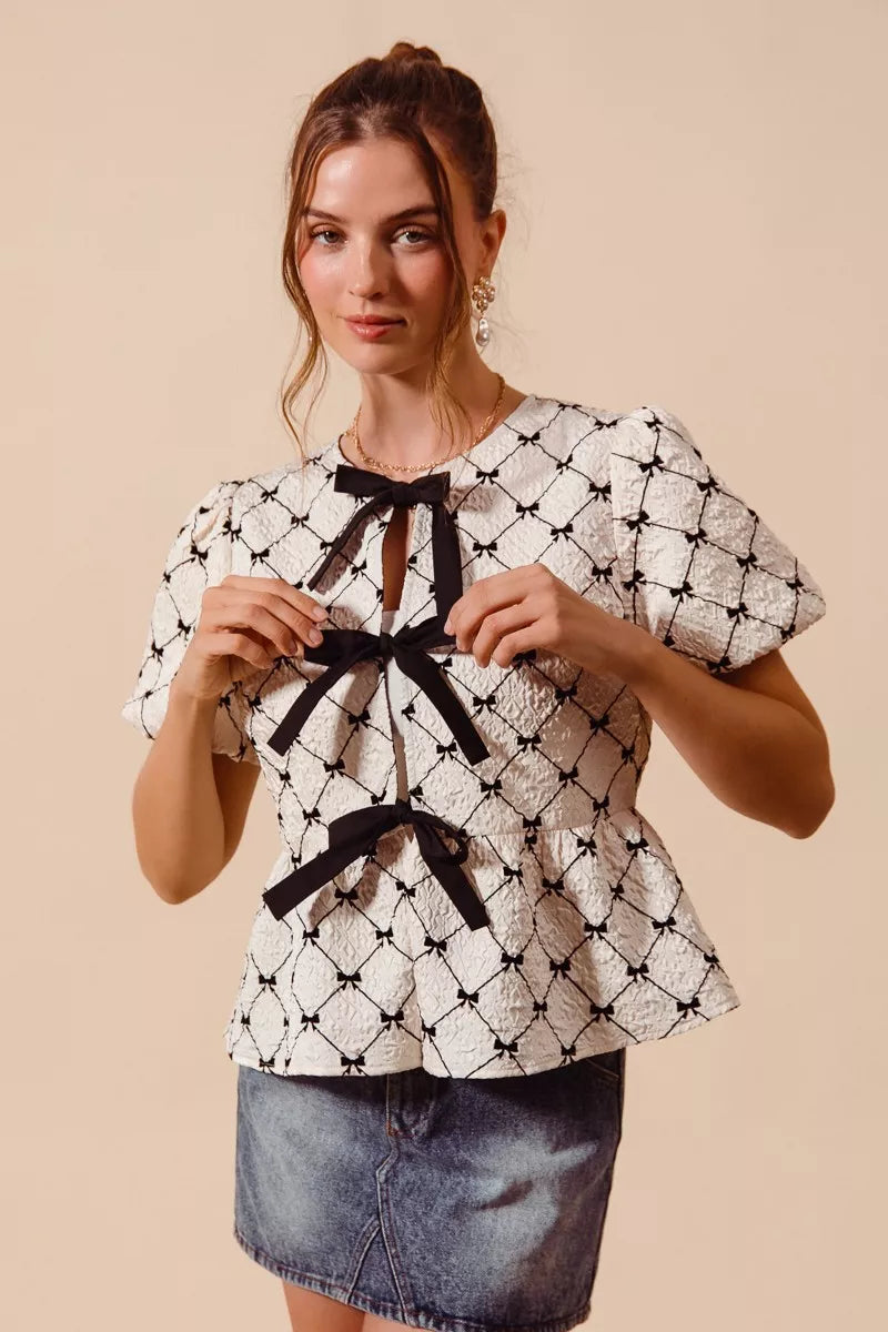 Ivory woven peplum top with black bow tie at neckline and puff sleeves, centered front.