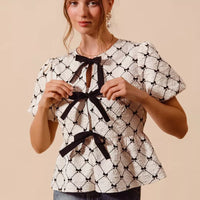 Ivory woven peplum top with black bow tie at neckline and puff sleeves, centered front.