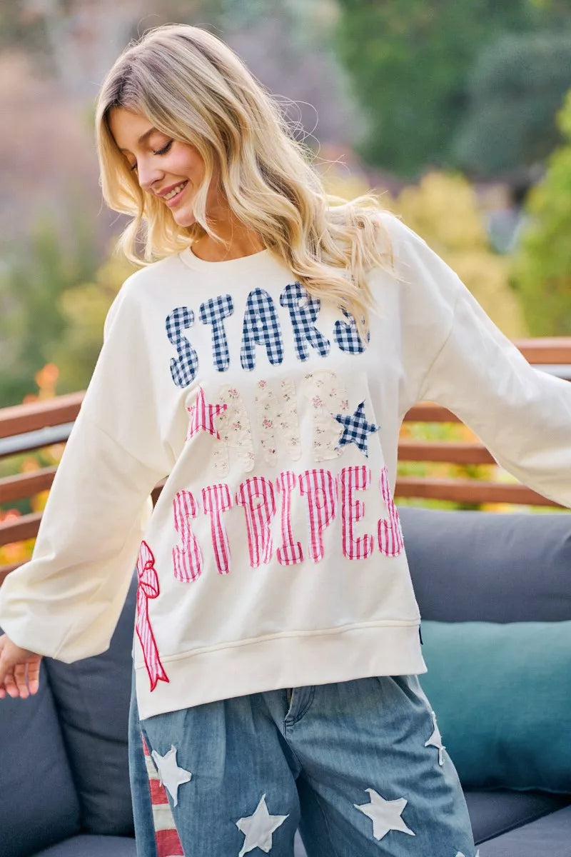 Cream sweatshirt featuring star-and-stripe patch lettering with red and blue bow details.