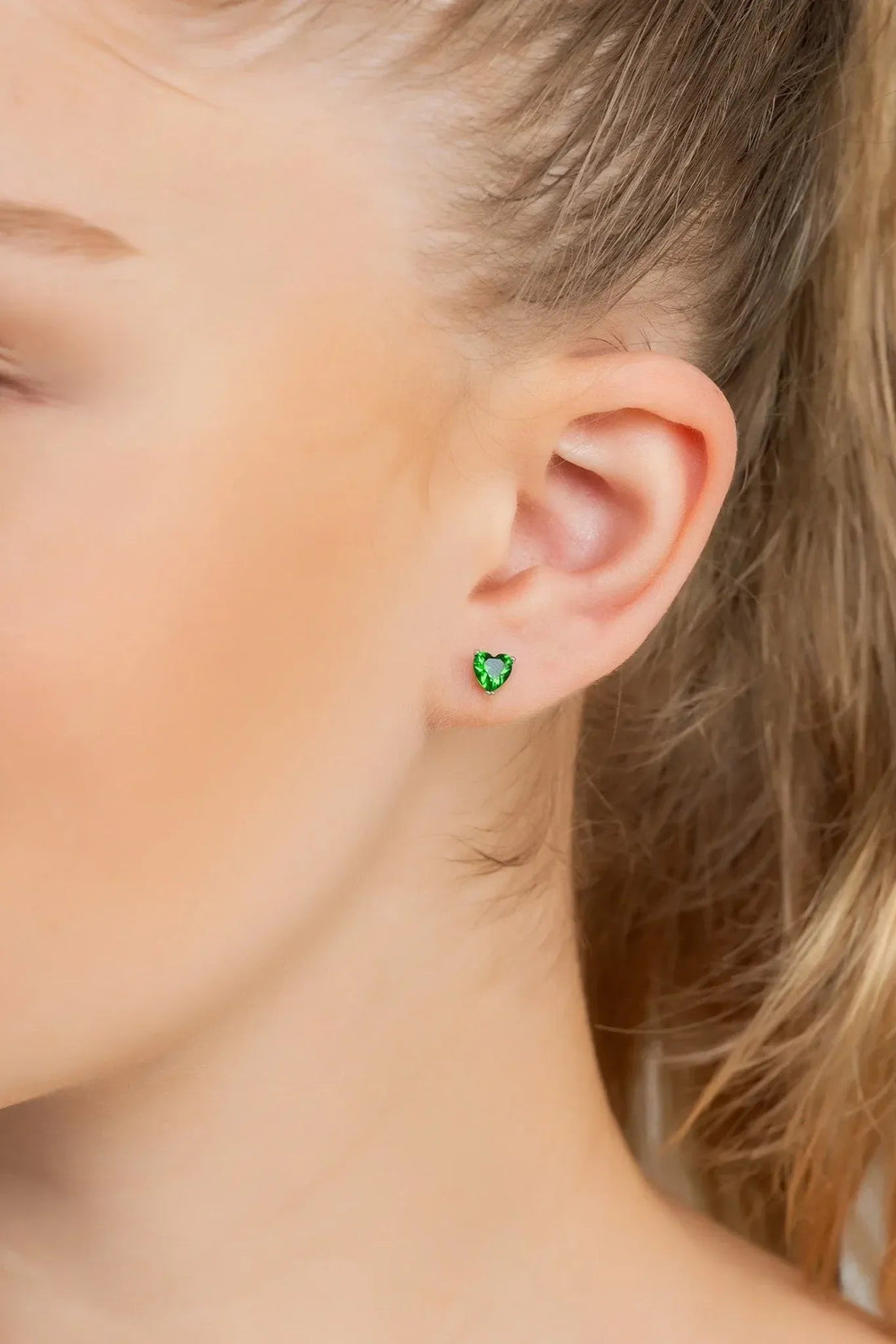 Emerald-green heart stud earrings on ear with small silver posts