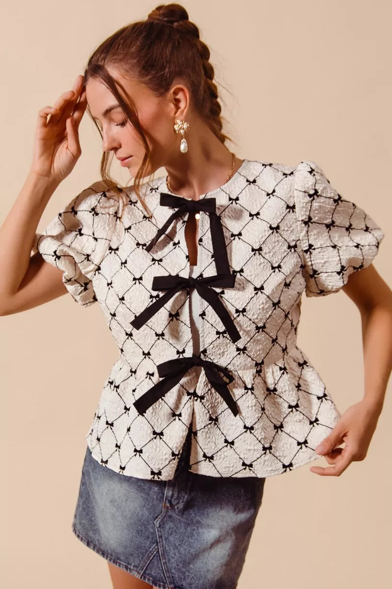 Ivory woven peplum top with black bow tie at neckline and puff sleeves, angled side view.