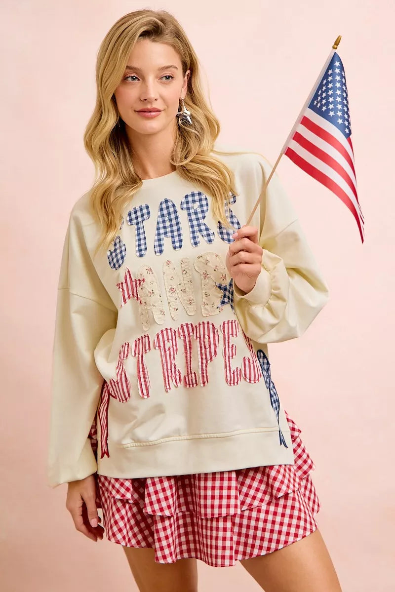 Cream sweatshirt with star-and-stripe patch graphic and red-blue bow detail on sleeve.
