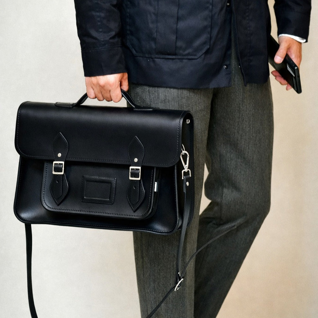 Black leather satchel carried by a person with front buckle flap and shoulder strap.