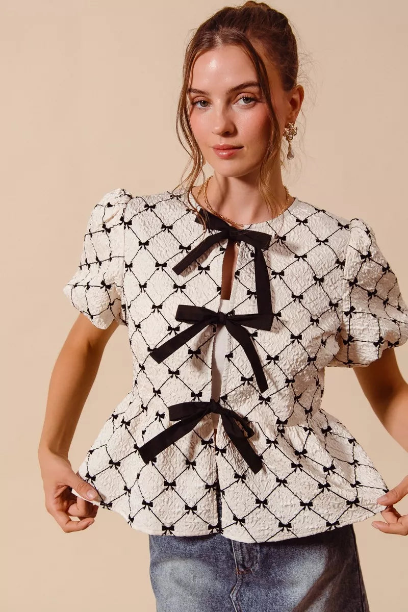 Ivory woven peplum top with black ribbon bows and puff sleeves, close front view.
