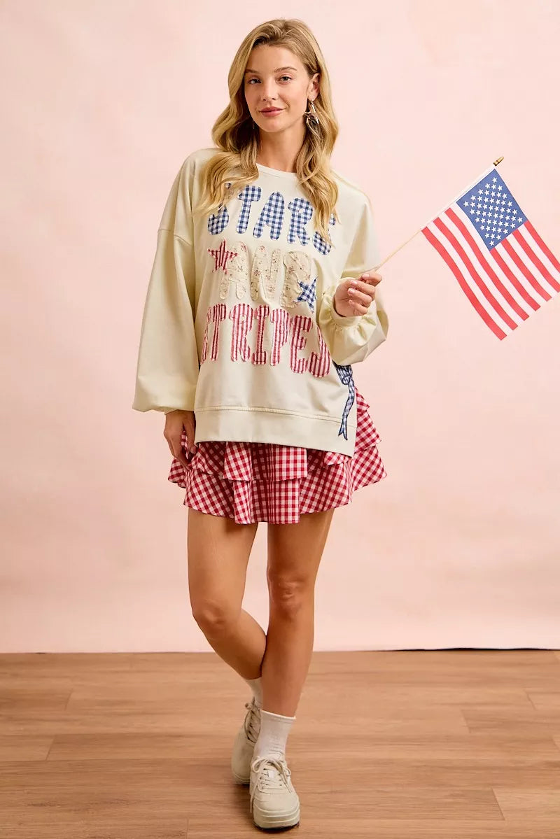 Cream sweatshirt with star-and-stripe patch graphic and blue bow detail near lower hem.