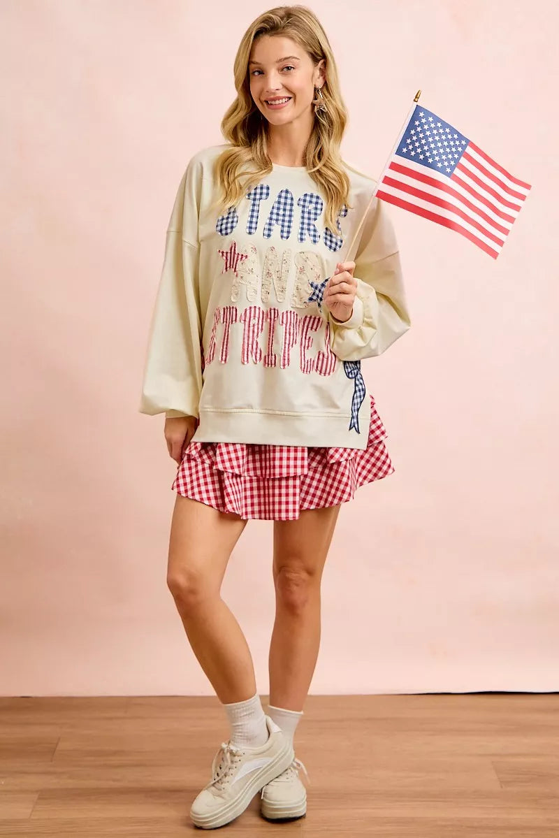 Cream sweatshirt with stars-and-stripes graphic, patch-style print, long sleeves; standing pose