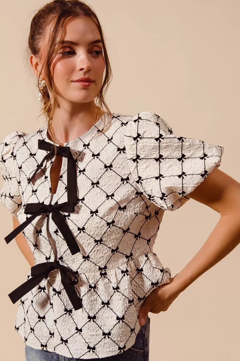 Ivory woven peplum top close-up with black bow ties at neckline and lattice print.