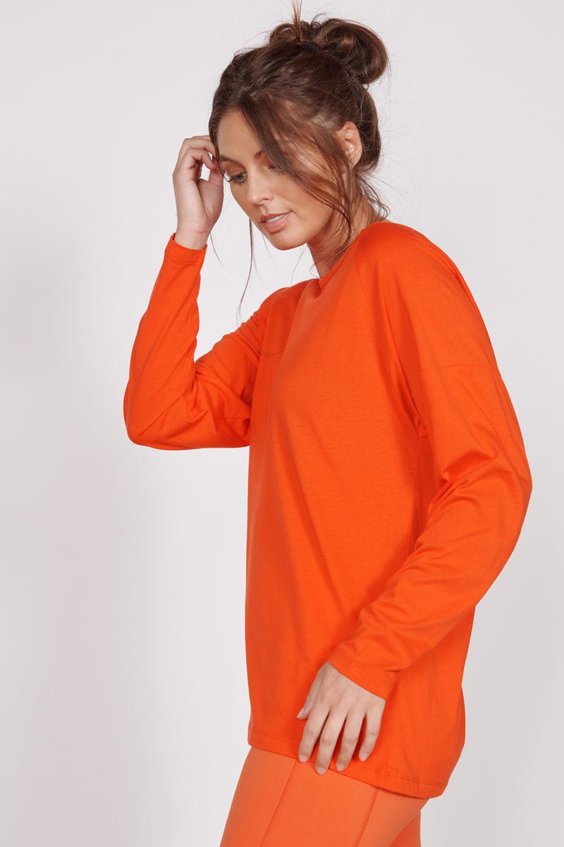 Orange long-sleeve training top, side view with draped hem and relaxed fit